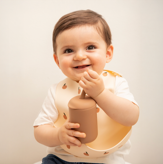 Bebé usando pechera de silicona y vaso de aprendizaje en tonos tierra, ideal para alimentación complementaria